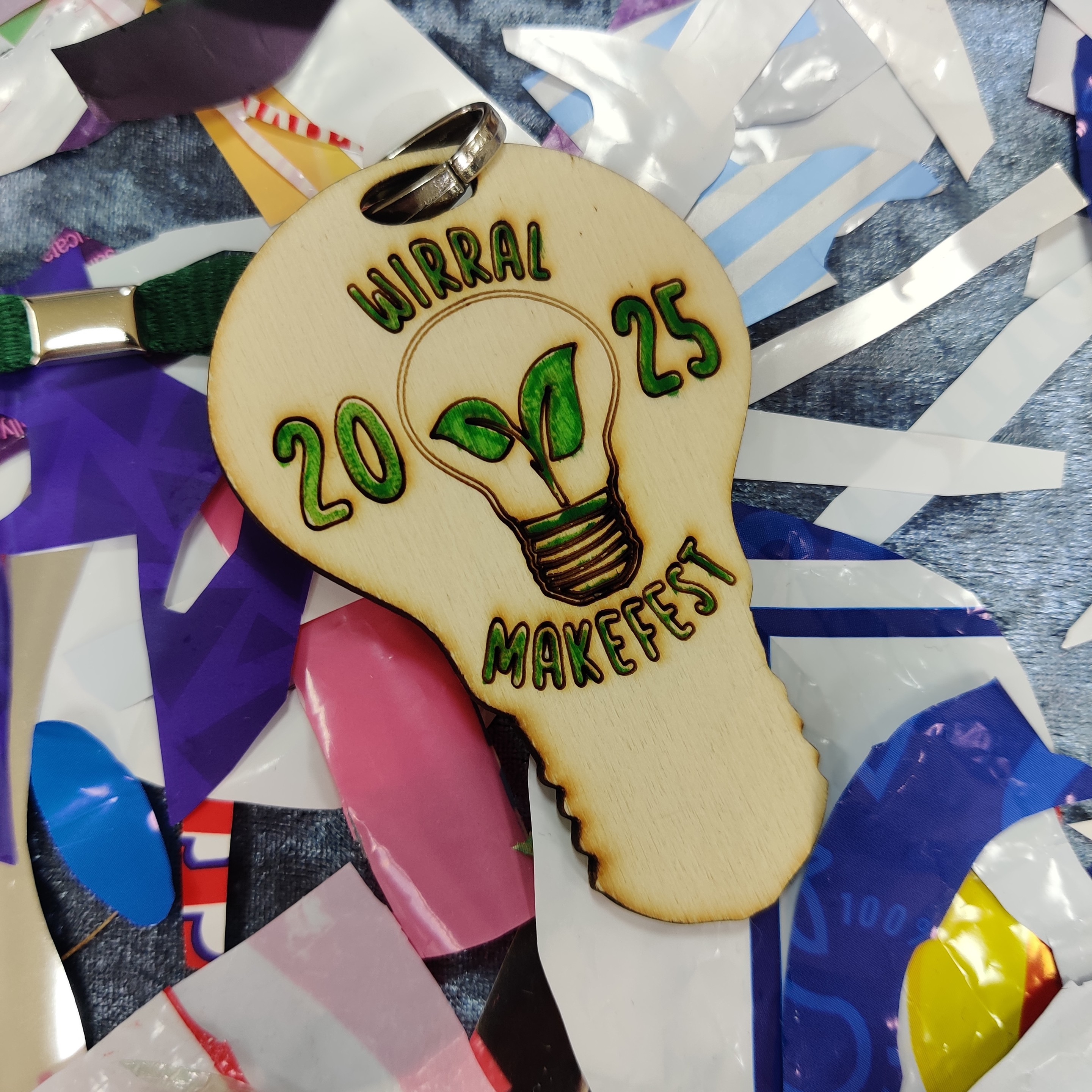 Laser cut Wirral MakeFest lanyard in the shape of a lightbulb, on a background of plastic scraps