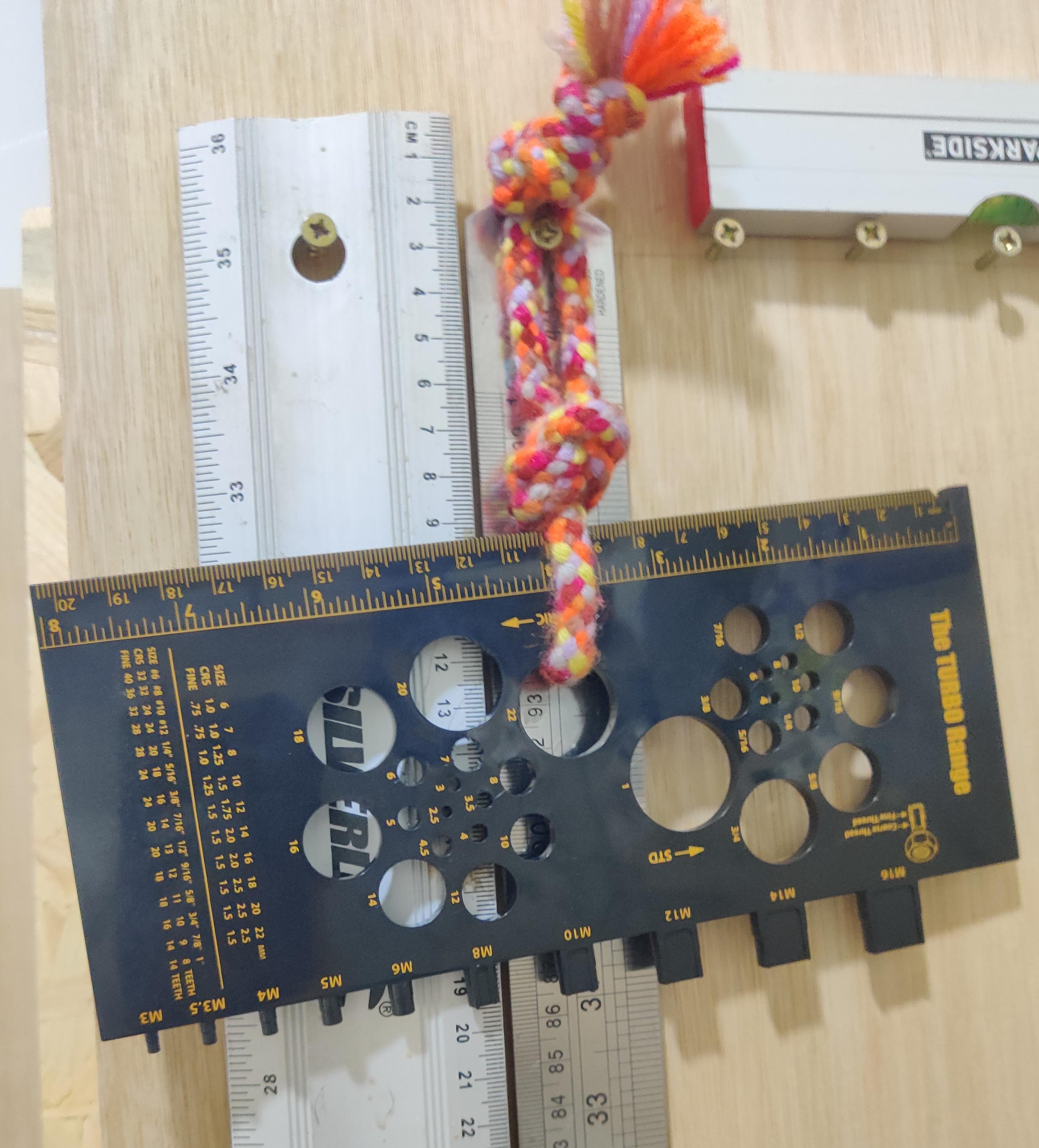 Black screw measuring gauge with gold lettering, with steel rules, hanging from screws attached to a wall.