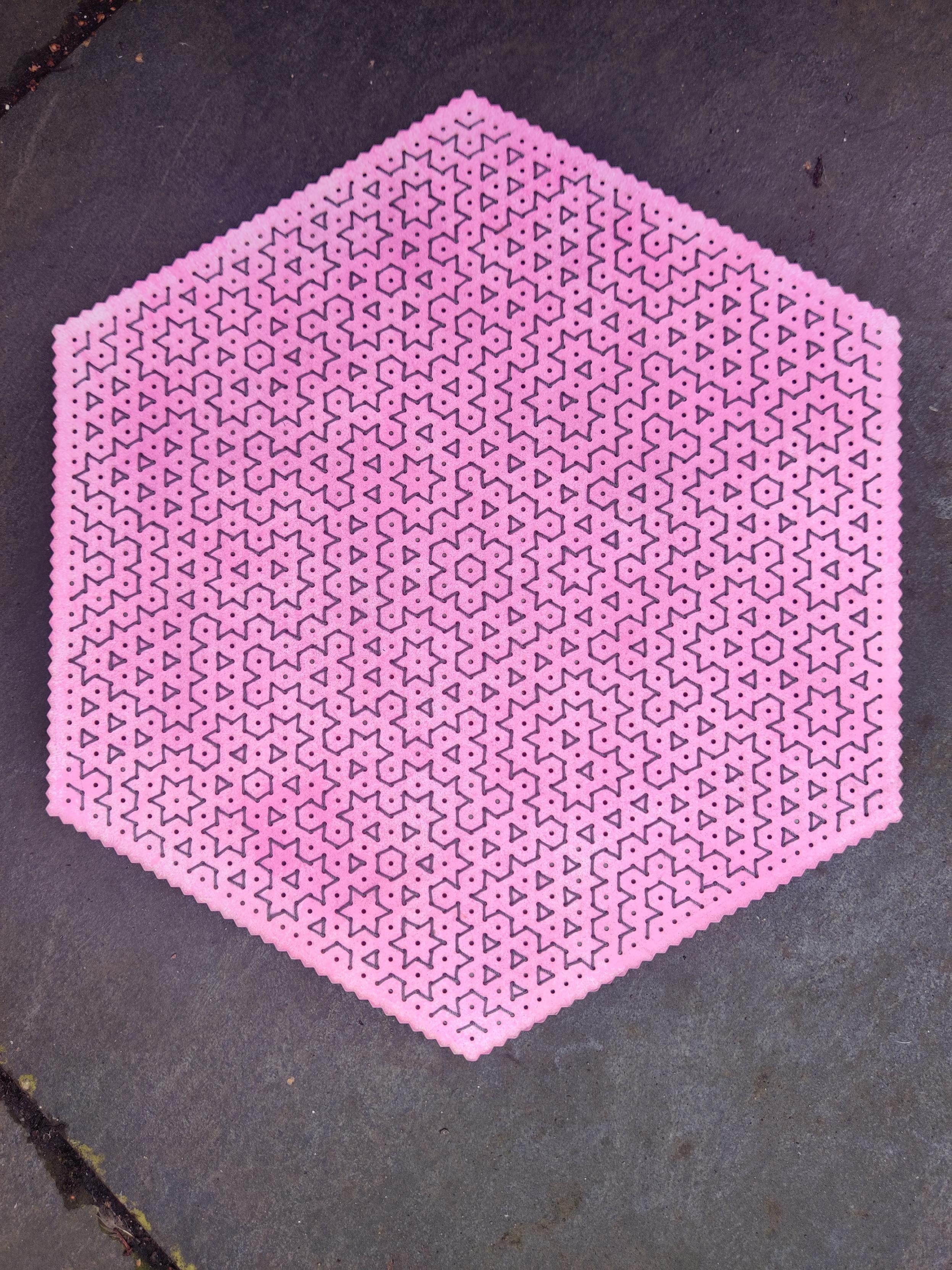 Hitomezashi embroidery in dark grey crochet cotton on a hexagonal piece of card which has been unevenly coloured with a pink stamp and laser cut with an isometric dot grid and zigzag edges