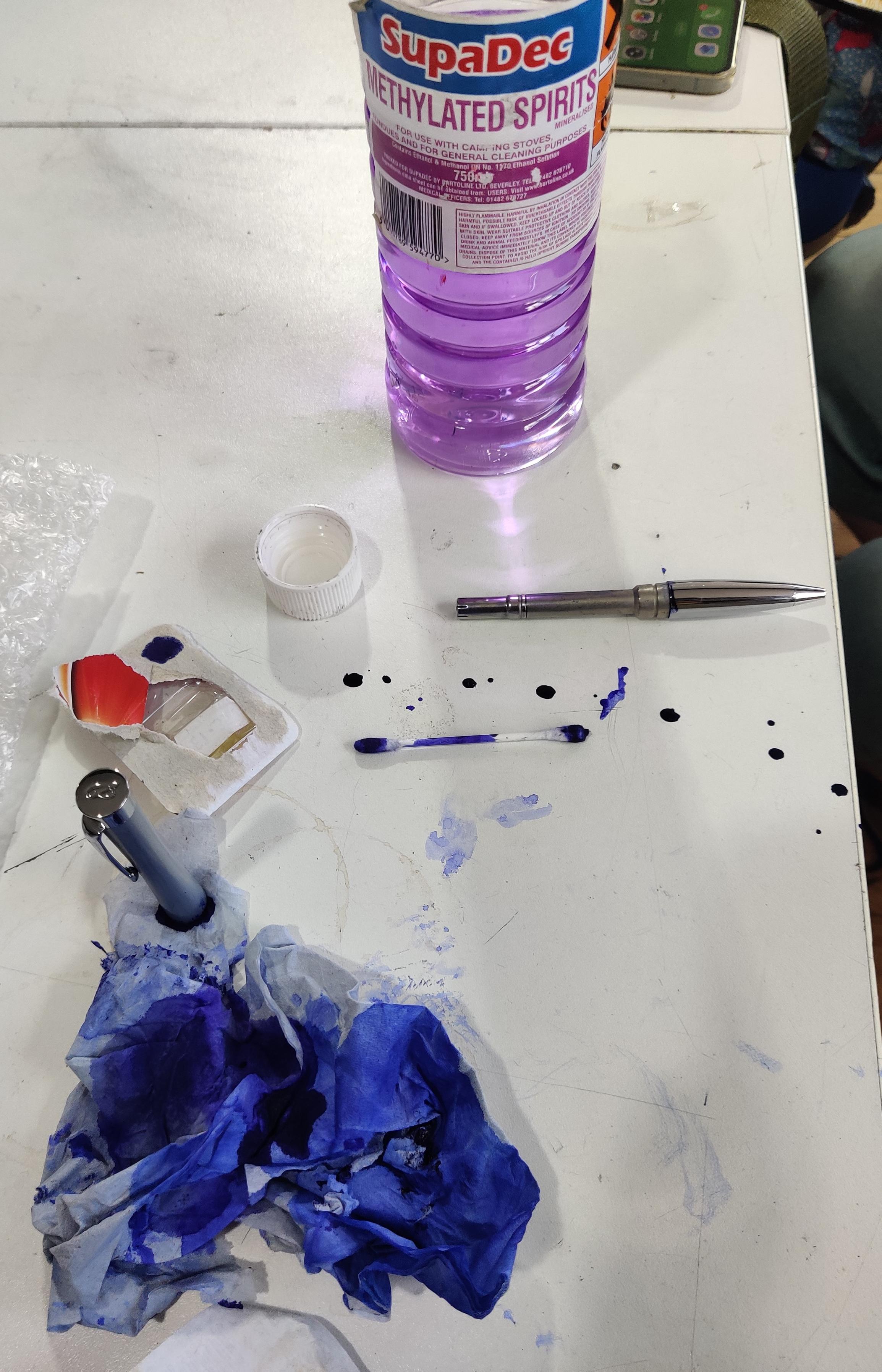 A white table with inky rags, a bottle of meths and a silver pen