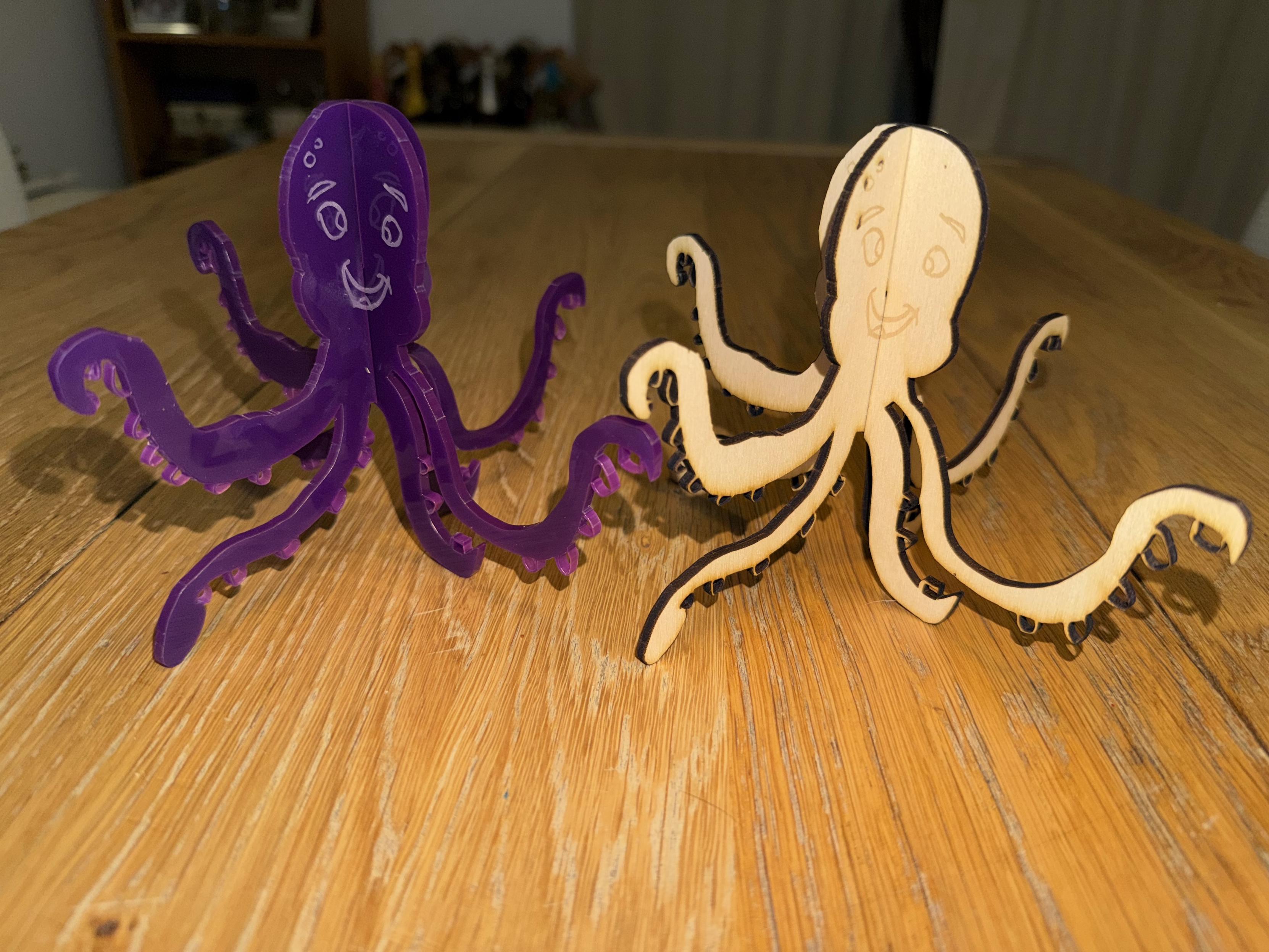 Two cartoonish octopus figures are placed on a wooden table. One is made of purple plastic, and the other is crafted from light-colored wood, both featuring smiling faces and playful designs. They are made from laser cut materials, interlocked at 90 degrees. 