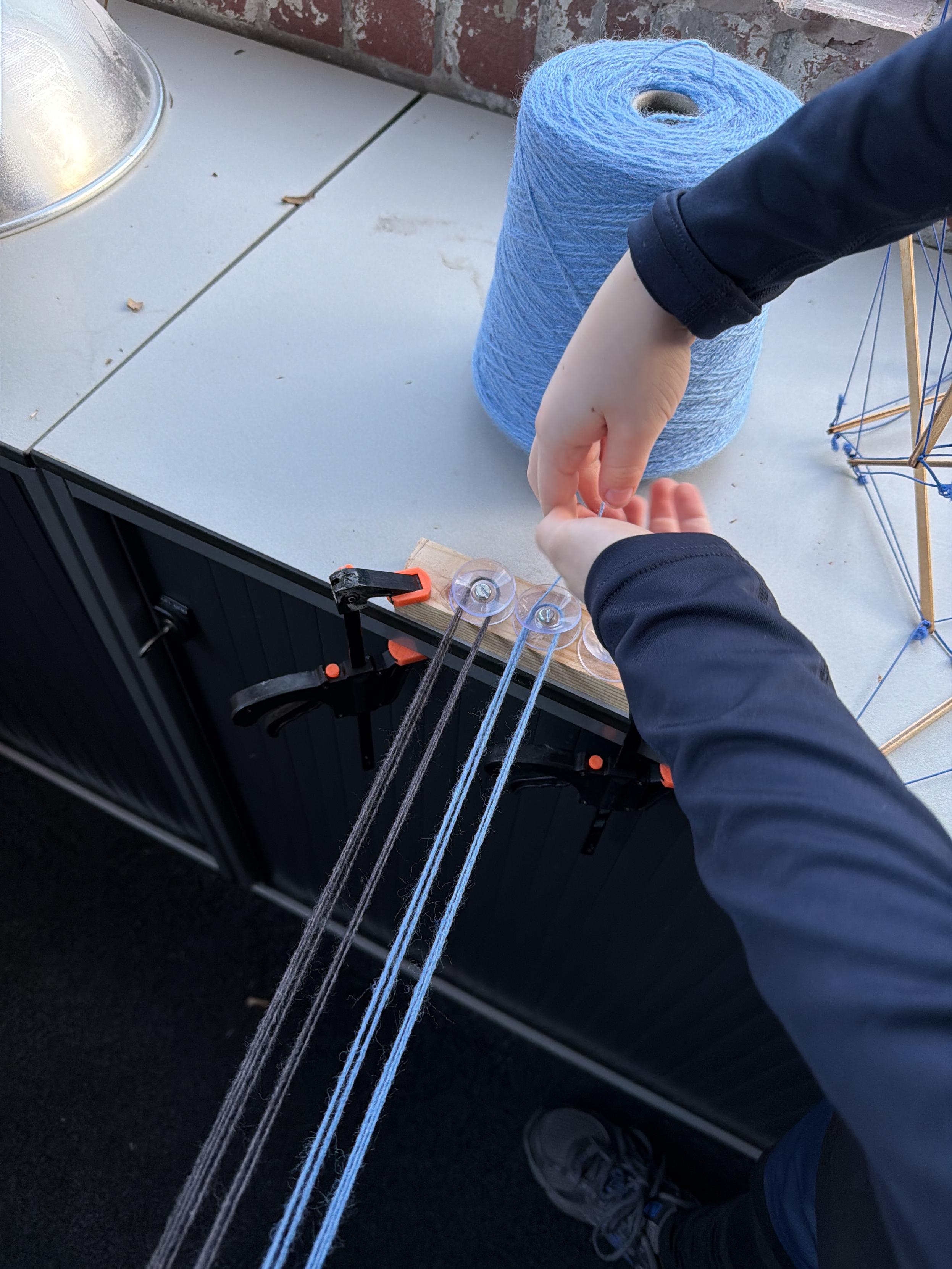 A close-up of a hand working with yarn on a table. A spool of light blue yarn is visible alongside a wooden board with clamps that holds several strands of yarn, including blue and dark threads. The setting appears to be indoors with a brick wall