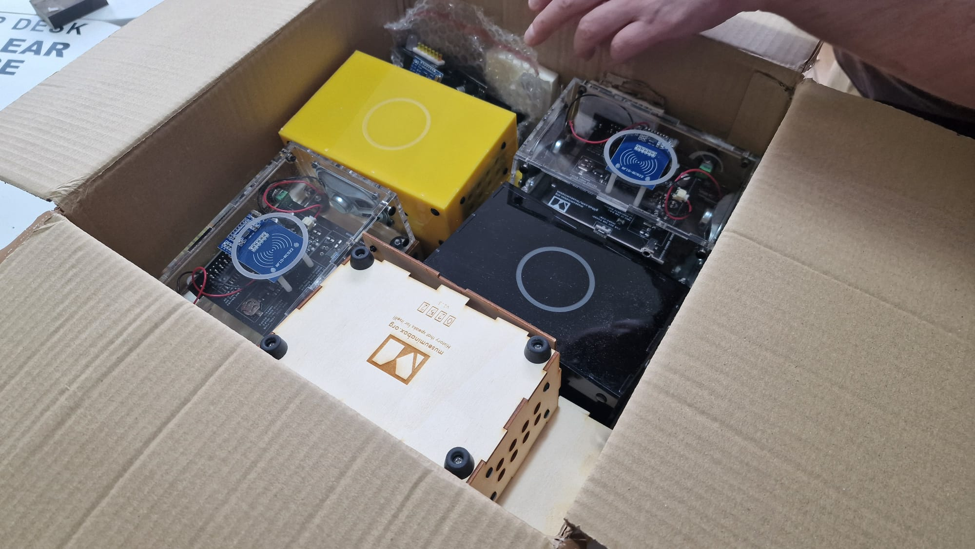 An open cardboard box showing a collection of Museum in a Box boxes inside: a couple of transparent ones, a yellow one, a black one and a wooden one. The front of the wooden one can just be seen to not be connected to the rest of the box...