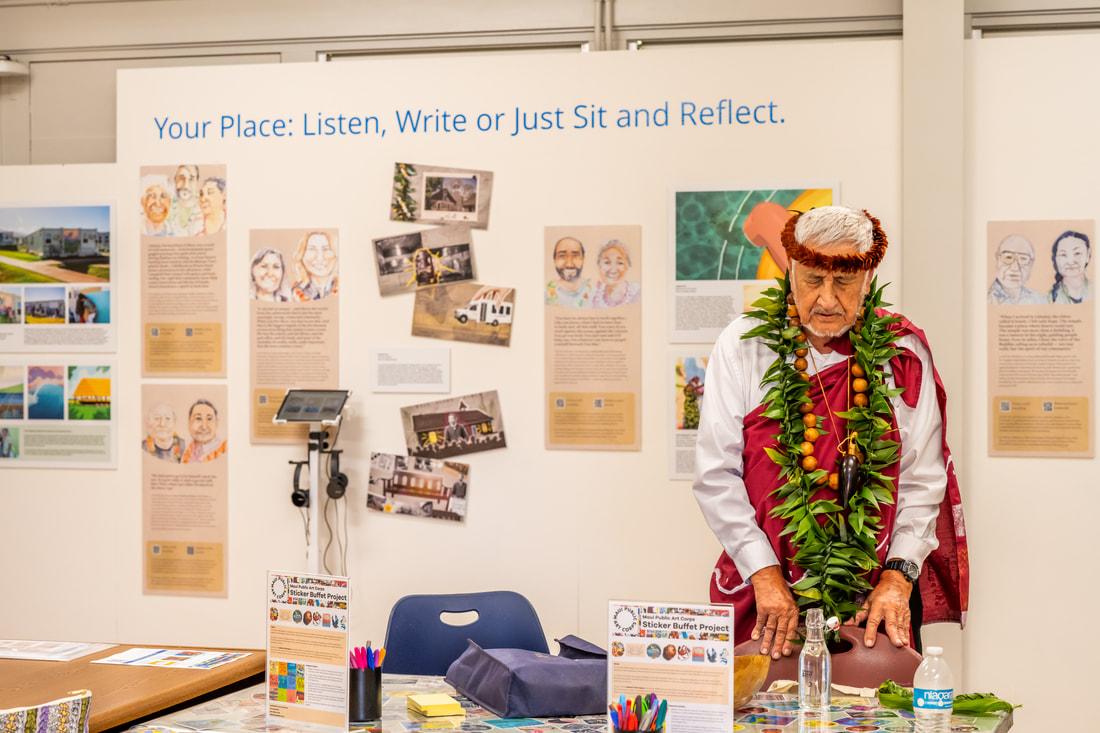 Opening the space with a traditional blessing led by Uncle Bill Garcia – Photo courtesy County of Maui A view of the exhibition being blessed, on the wall in the background are pictures on the contributors to case studies and the phrase “Your place: Listen, Write or Just Sit and Reflect”