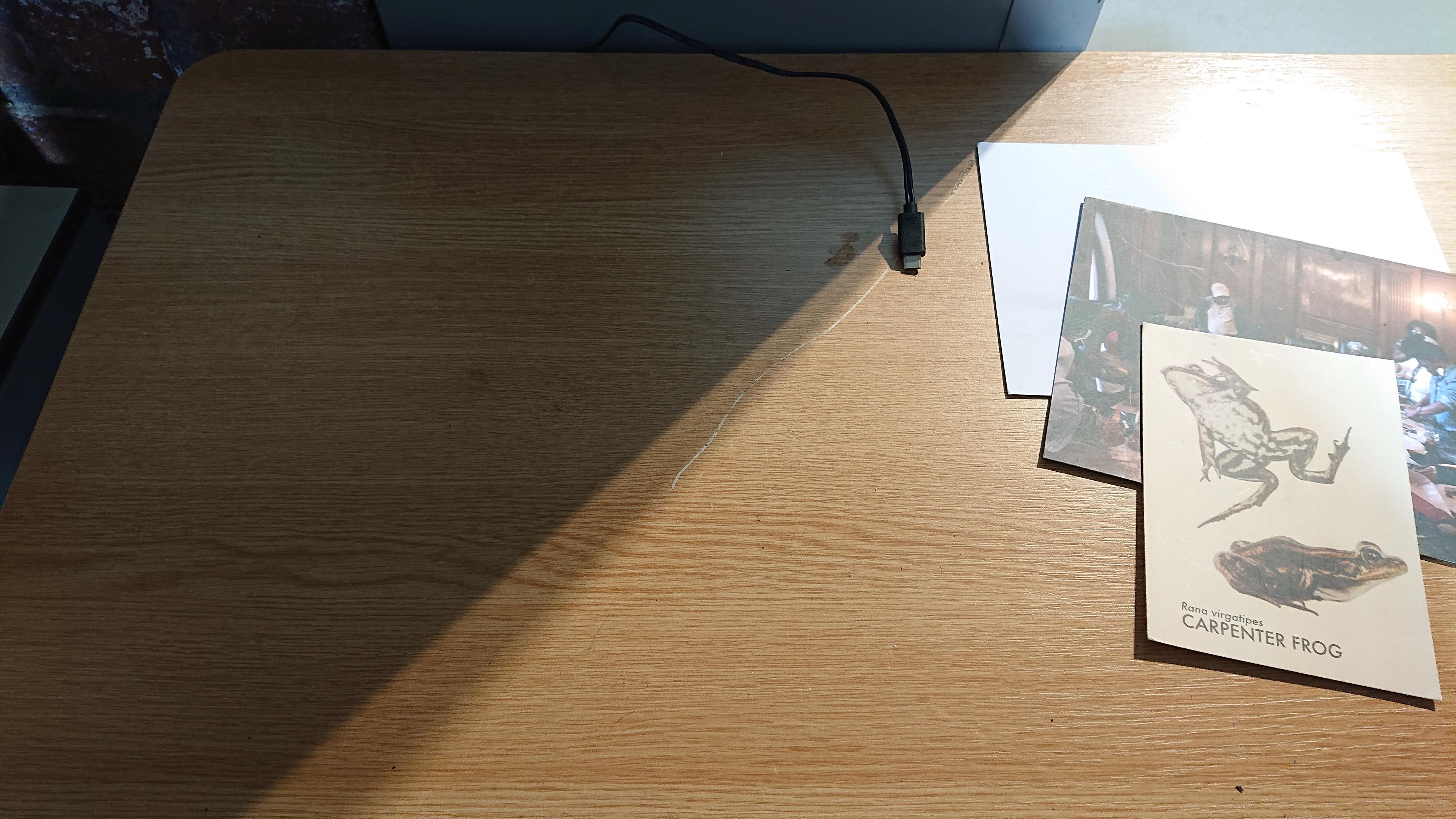 A table with a couple of picture postcards off to the side and a lonely USB-C cable poking over the edge at the back. The focus is on an empty area next to both things, about the size of a Museum in a Box