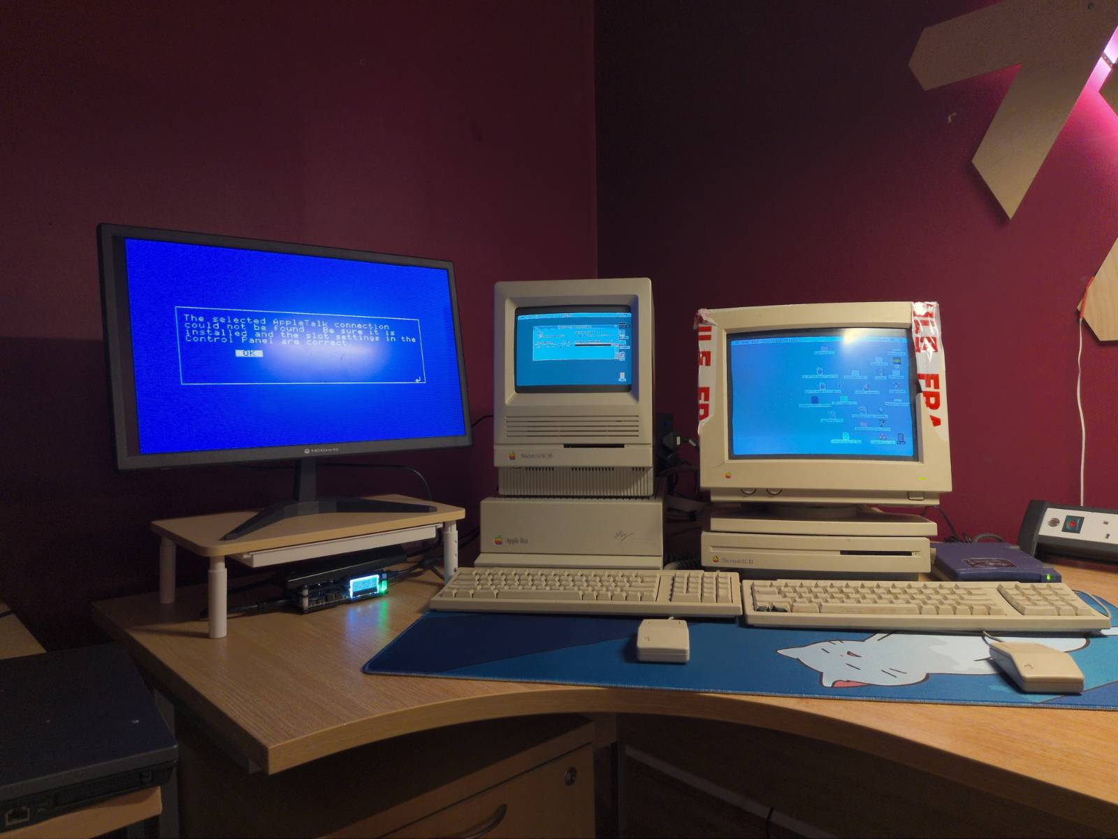 A retro computer setup featuring two beige Macintosh computers with CRT monitors, a modern monitor displaying pixelated text, and a cute cat-themed mousepad. The room has purple walls and warm lighting. A glowing Nix logo is featured in the background.